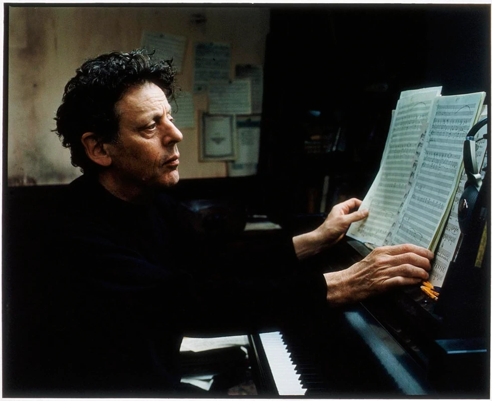 Philip Glass in black and white sitting at the piano, looking at a music score, evoking the composition process of Koyaanisqatsi and the relationship between music, rhythm, and movement that inspires the sound of modern technology.